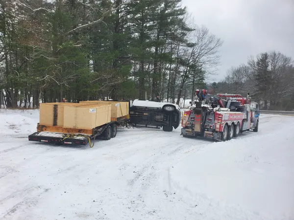 Heavy wrecker beside a rolled-over pickup and loaded trailer in the snow