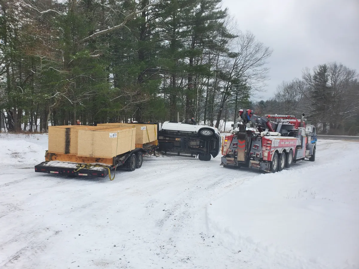 Heavy wrecker setting up beside a rolled-over pickup and loaded trailer in the snow